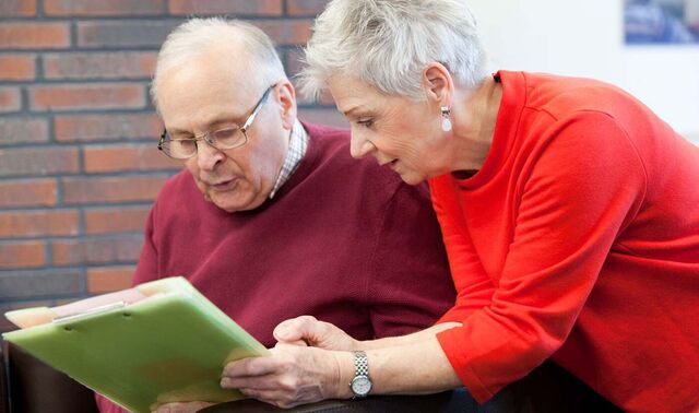 Eine Dame ist zu einem Senioren hintergebeugt, welcher etwas liest.