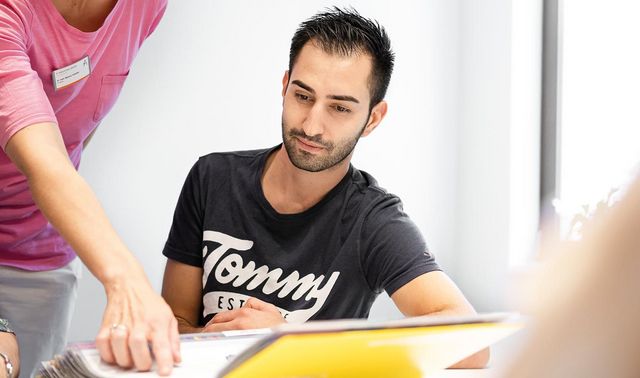 Ein junger Mann sitzt vor einer Akte an einem Tisch. Eine Frau, die neben ihm steht zeigt auf etwas in der Akte.
