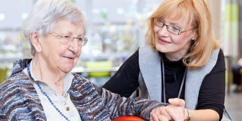 Eine Frau sitzt neben einer Seniorin und hält ihre Hand während sie ihr zugewandt ist.