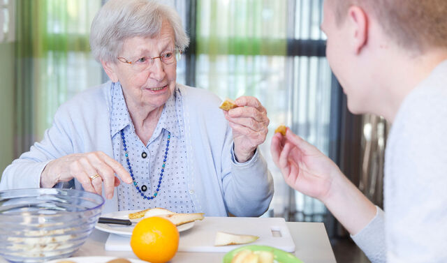 Eine Seniorin und ein junger Mann sitzen an einem Tisch und essen gemeinsam ein Stück Birne.