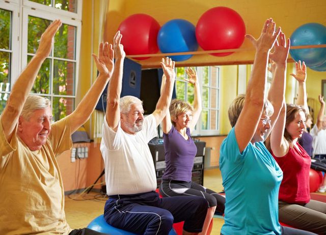 Einige Senioren sind in einem Trainingsraum und sitzen auf Gymnastikbällen, während sie ihre Arme nach oben gerichtet haben.