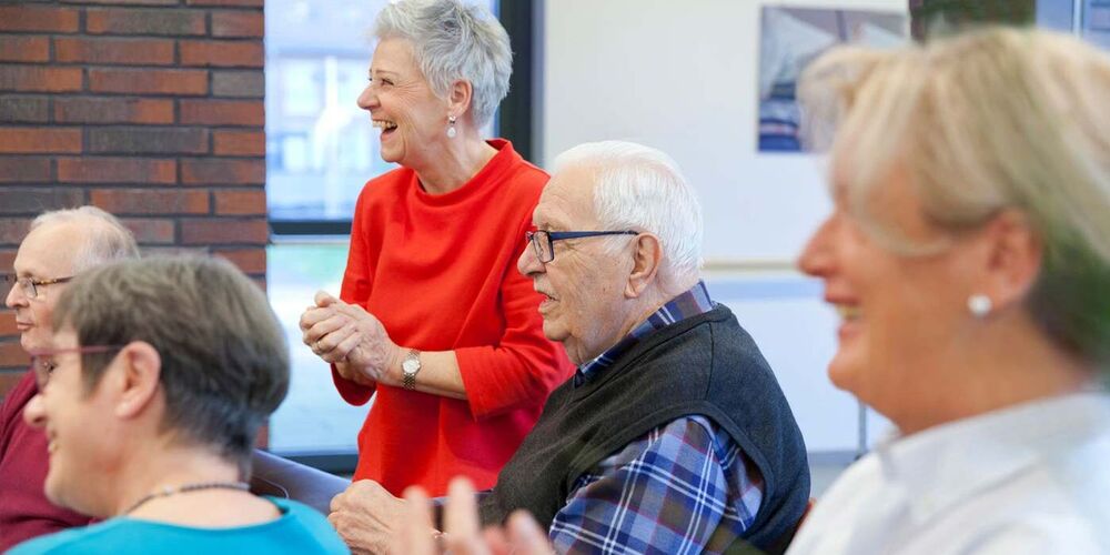Mehrere Seniorinnen und Senioren sitzen in einem Kreis und singen und klatschen. Eine Ehrenamtliche steht bei ihnen und klatscht ebenfalls.